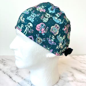 May include: A dark teal scrub cap featuring a repeating pattern of pastel-colored creatures including aliens, yetis, and rabbits with button eyes. The fabric design incorporates small stars and various whimsical characters in shades of pink, blue, and green.