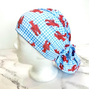 May include: A blue and white gingham patterned scrub cap featuring red lobster illustrations. The cap is tied in the back and is displayed on a mannequin head.