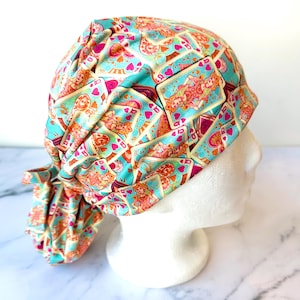 May include: A colorful scrub cap featuring a repeating pattern of playing cards in shades of teal, orange, pink, and cream. The cap is tied in the back and is displayed on a white mannequin head.