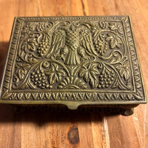 May include: A small, rectangular brass box with an ornate design. The box features a raised eagle with grapevines and leaves surrounding it. The box has a hinged lid and sits on four small feet.