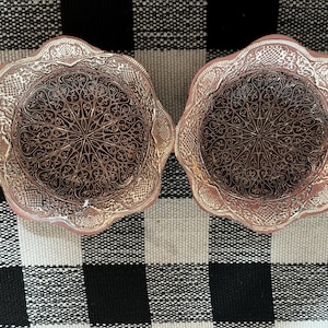 May include: Two decorative, scalloped-edge bowls with intricate, symmetrical patterns. The bowls have a metallic, rose-gold finish with a darker, patterned interior. The bowls are displayed on a black and white checkered surface.