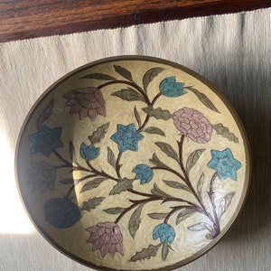 May include: A brass bowl with a floral design. The bowl is decorated with blue, green, and pink flowers on a cream background.