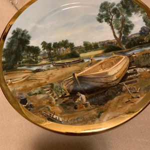 May include: A decorative ceramic plate with a gold rim features a painted scene of a boat being built on a riverbank. The scene includes trees, a river, and people working on the boat.