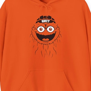 Op de afbeelding: Oranje hoodie met een cartoonfiguur met grote ogen en een brede grijns. Het personage draagt een zwarte hoed met het woord "GRIT". De hoodie heeft een voorzak en een capuchon met trekkoord. De mouw heeft het woord "Hustle".