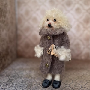May include: A handcrafted poodle figurine wearing a gray coat with gold buttons, a gray scarf, and black shoes. The poodle holds a small paper in its paw. The poodle has white fur and a black nose.