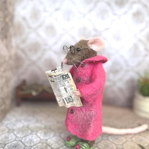 May include: A handmade felt mouse wearing a pink coat, glasses, and green shoes, reading a newspaper. The mouse has brown fur, a pink ear, and a long tail. The miniature scene includes a small bed and a plant.