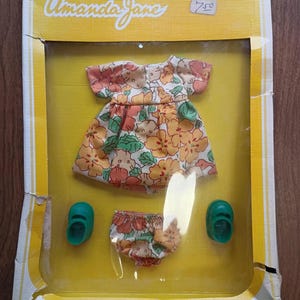 May include: A vintage doll clothing set, featuring a floral print dress, matching bloomers, and two green shoes. The packaging is yellow with the name "Amanda Jane" in cursive.
