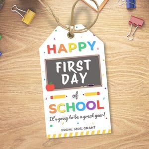 May include: A white tag with the words "HAPPY FIRST DAY of SCHOOL" in colorful letters. A small chalkboard illustration is in the center, with an apple and pencil graphics. The tag reads "It's going to be a great year!" and "FROM: MRS. GRANT."