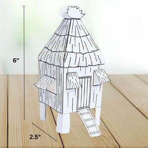 May include: A black and white paper model of a thatched-roof hut, approximately 6 inches tall and 2.5 inches wide. The hut features a door, a ladder, and two small awnings. The model is set on a wooden surface.
