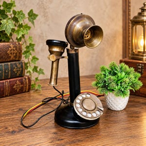 Könnte beinhalten: Ein Telefon im Vintage-Stil mit einem schwarzen zylindrischen Sockel und Schaft, einer Wählscheibe aus Messing sowie einem Hörer und Mundstück aus Messing. Das Gerät verfügt über ein schwarzes Kabel mit gelben und roten Akzenten am Sockel.