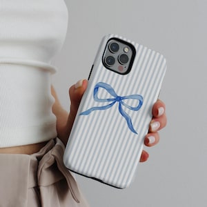 May include: A smartphone case featuring a light blue and white vertical striped pattern with a centered watercolor-style blue ribbon bow graphic. The case has a black camera lens cutout area and covers the back of the mobile device completely.