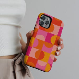 May include: A colorful phone case with a geometric pattern. The case features a repeating design of pink, orange, and yellow shapes, including squares and semi-circles. The case is held in a person's hand.