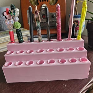 May include: A tiered, light pink pen holder with multiple pens and styluses. The holder has three levels, each with several circular openings to hold pens. Pens include black, pink, and glittery options. A decorative pen with a butterfly design is also visible.