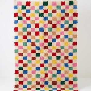 May include: A rectangular area rug featuring a repeating checkerboard pattern of small squares in vibrant colors including red, blue, yellow, green, pink, and tan against a cream-colored background, finished with a short fringe at the bottom edge.