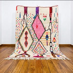 May include: A rectangular area rug with an off-white base featuring a large central pink diamond shape, a smaller diamond with a multicoloured grid pattern, and various geometric motifs in red, blue, yellow, and purple, finished with a fringed edge.