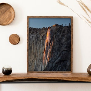 May include: A framed photograph of a waterfall cascading down a rocky cliff face. The waterfall is illuminated by the setting sun, creating a warm orange glow. The photograph is displayed on a wooden shelf with other decorative items.