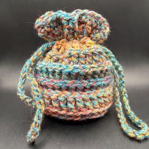 May include: A small, round, drawstring pouch crafted from variegated yarn in shades of blue, yellow, orange, and pink. The crochet construction features a textured, ribbed pattern throughout the body and a cinched top with two long, braided yarn ties.