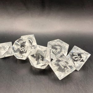 May include: A set of clear, transparent polyhedral dice for tabletop games. The dice are made of a clear material and feature etched numbers and symbols. The set includes various shapes, such as a d4, d6, d8, d10, d12, and d20.