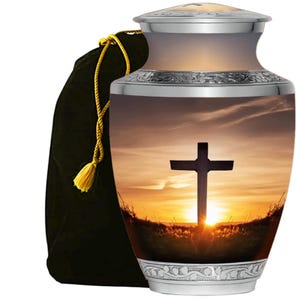 May include: A silver and beige cremation urn with a sunset scene featuring a cross. The urn has a decorative silver band around the top and bottom. A black velvet bag with a gold tassel is on the left.