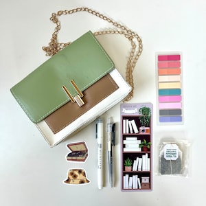Puede incluir: Un bolso cruzado verde, marrón y blanco con una cadena dorada, bolígrafos, pegatinas, un marcapáginas y té. El marcapáginas dice "Books of" y representa una estantería. Las pegatinas incluyen un tocadiscos y un sombrero de sol.
