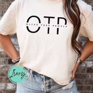 May include: Cream-colored t-shirt with the text "OTP OLIVE TREE PEOPLE" in black. The shirt is a casual style, with short sleeves and a relaxed fit. The person is wearing blue denim shorts.