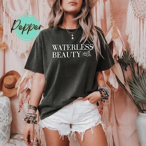 May include: Dark gray t-shirt with the text "WATERLESS BEAUTY" in white, accompanied by a floral design. The shirt is worn with white shorts. The word "Pepper" is in a teal circle in the upper left corner.