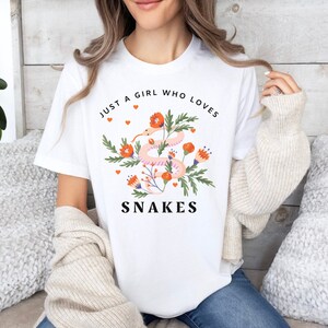 May include: White t-shirt with a floral design and a pink snake. The text "Just a girl who loves snakes" is printed on the shirt.
