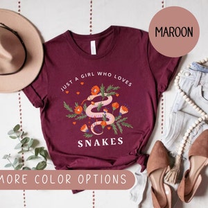 May include: A maroon t-shirt with the text "Just a girl who loves snakes" and a graphic of a snake with flowers. The shirt is laid flat on a white surface with a hat, jeans, and shoes.