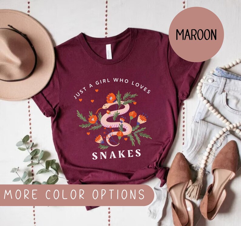 May include: A maroon t-shirt with the text "Just a girl who loves snakes" and a graphic of a snake with flowers. The shirt is laid flat on a white surface with a hat, jeans, and shoes.