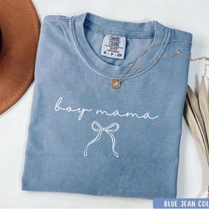 May include: A light blue t-shirt with the text "boy mama" in white script with a white bow underneath. The shirt is made of a soft, comfortable material.