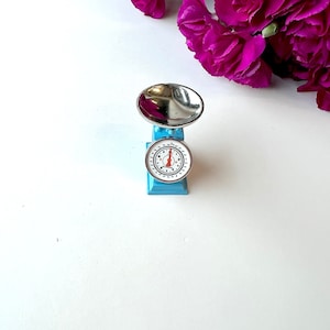 Op de afbeelding: Een kleine blauwe speelgoedkeukenweegschaal met een zilveren kom en een witte wijzerplaat met zwarte cijfers. De weegschaal staat op een wit oppervlak.