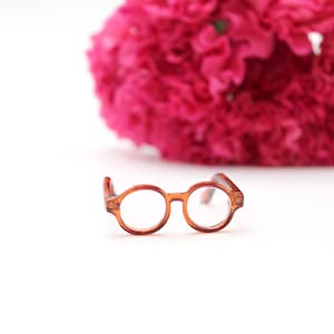 May include: Small, round, amber-colored eyeglasses with clear lenses are centered in the frame. A large, out-of-focus, pink flower is in the background. The glasses have a classic, vintage style.