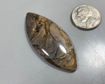 May include: A polished, light brown, gray, and black agate cabochon with a marbled pattern. The cabochon is shaped like a teardrop and is approximately 2 inches long.