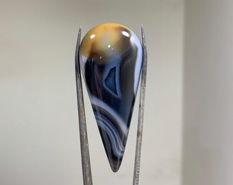 May include: A teardrop-shaped agate cabochon with a white, brown, and black banded pattern. The cabochon is held in place by two metal tweezers.