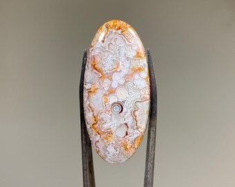 May include: A white and orange agate cabochon with a swirling pattern. The cabochon is oval shaped and has a smooth, polished surface.