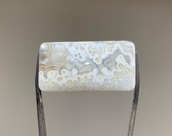 May include: A white agate cabochon with a swirling pattern of white, gray, and brown. The cabochon is rectangular and has a smooth, polished surface.