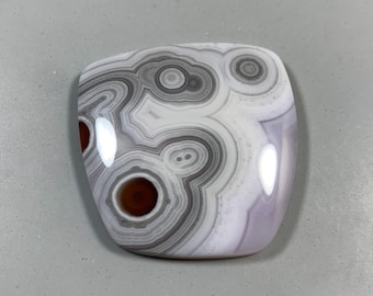 May include: A polished, rounded stone with a unique pattern. The stone features concentric circles in shades of gray, white, and brown, creating an eye-catching design. The stone's surface is smooth and reflective, with a slight purple hue.