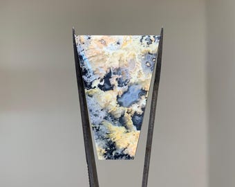 May include: A triangular shaped, polished agate cabochon with a black, white, and yellow pattern. The cabochon is held in place by two metal prongs.