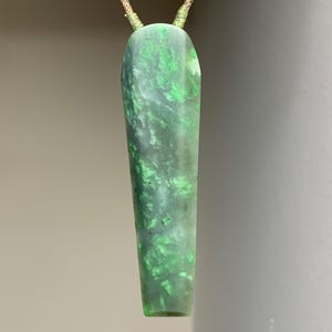 May include: A green and gray pendant with a rectangular shape and rounded top. The pendant has a marbled appearance with lighter green accents. It is suspended from a thin, tan cord.