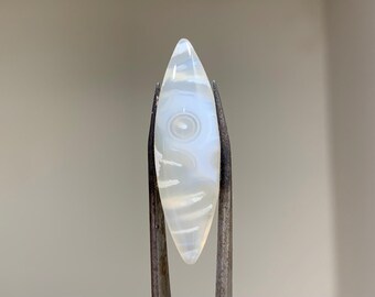 May include: White agate cabochon with a swirling pattern, shaped like a teardrop, held by tweezers.