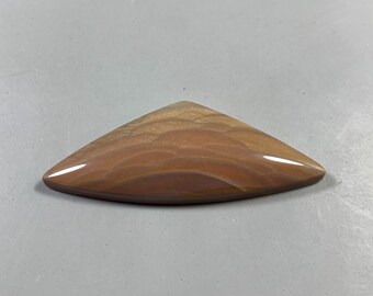 May include: A polished, triangular gemstone with a warm color palette of browns and tans. The stone has a smooth, glossy surface and a unique, layered pattern, resembling a landscape. The shape is reminiscent of a stylized arrowhead or a teardrop.