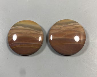 May include: Two round, polished stones with a layered, earthy color pattern of brown, tan, and cream. The stones have a glossy finish and a smooth, rounded shape, suggesting they are decorative or jewelry components.