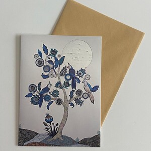 May include: A greeting card featuring a tree with blue birds and floral accents, set against a full moon. The card is paired with a brown envelope, all on a white surface. The artwork has a vintage, illustrated style.