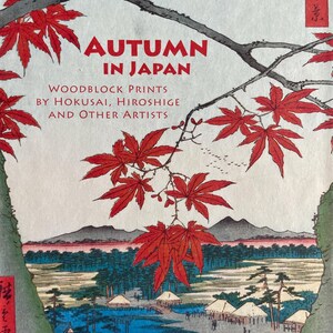 May include: A book cover featuring a Japanese woodblock print with the title "Autumn in Japan." The image showcases vibrant red maple leaves, a serene landscape with mountains, water, and traditional buildings. The text includes the names of artists Hokusai and Hiroshige.
