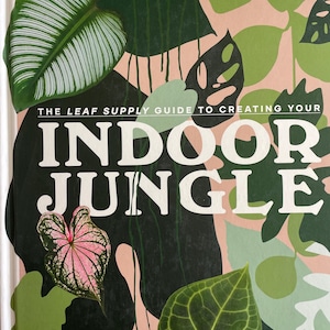 May include: A book cover with the title "INDOOR JUNGLE" in large white letters. The cover features a collage of green, pink, and white leaves against a light pink background. The text "THE LEAF SUPPLY GUIDE TO CREATING YOUR" is above the title. The authors' names are at the bottom.