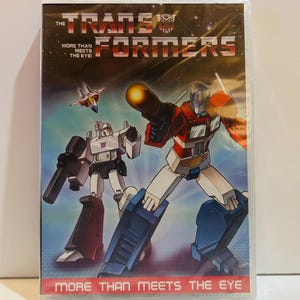 Op de afbeelding: Een dvd-hoes van de animatieserie Transformers. De cover toont Optimus Prime en Megatron in actie, met de slogan "More Than Meets The Eye". De achtergrond is een ruimtescène met een straaljager.