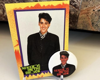 May include: A vintage New Kids on the Block trading card featuring a photo of a young man with dark hair and a black jacket. The card has a yellow, orange, and purple border with the band's name in green and black letters. A white button with the band's logo and name in red and black letters is also shown.