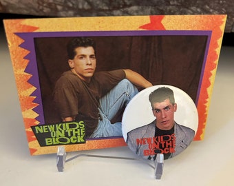 May include: A yellow and orange card with a photo of a young man with brown hair and a brown shirt. The card has the words "New Kids on the Block" printed on it. A white button with a photo of a young man with brown hair and a gray shirt is also pictured. The button has the words "New Kids on the Block" printed on it.
