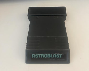 May include: A black Astroblast video game cartridge. The cartridge has the game title in teal lettering. The cartridge is rectangular with a ridged design on the sides. The cartridge is on a white surface.