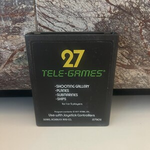 Puede incluir: Un cartucho de juego Atari negro con el número 27 y el texto "TELE-GAMES" impreso en amarillo. El cartucho enumera los juegos "SHOOTING GALLERY", "PLANES", "SUBMARINES" y "SHIPS". El texto "Program contents © 1977 ATARI, INC." y "Use with Joystick Controllers" también está impreso en el cartucho.